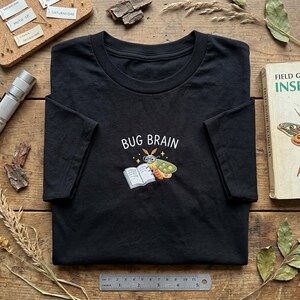 May include: Black t-shirt with the text "BUG BRAIN" above an embroidered design of a cartoon moth wearing glasses and reading a book. The shirt is on a wooden surface with a book and other items.