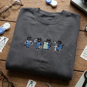 May include: A charcoal gray crewneck sweatshirt featuring an embroidered graphic of four black cats dressed in blue medical scrubs. The cats are depicted performing eye exams with tools including a trial frame, a handheld device, an eye chart, and a slit lamp.