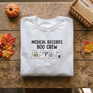 May include: A light gray crewneck sweatshirt featuring the black text MEDICAL RECORDS BOO CREW above a graphic of four white ghosts. One ghost holds a file folder, one holds a magnifying glass, one holds papers, and one holds a computer mouse.