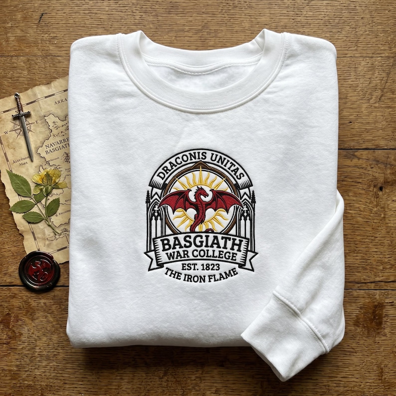 Puede incluir: Sudadera blanca con un emblema de Basgiath War College. El emblema presenta un drag&oacute;n rojo, un sol y texto, con las palabras "Draconis Unitas" y "The Iron Flame". La sudadera es de material suave.
