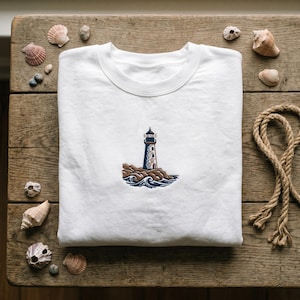 May include: A white crewneck sweatshirt featuring a small, centered embroidery of a lighthouse standing on brown rocks surrounded by dark blue stylized ocean waves. The design is positioned in the middle of the chest area on the plain white fabric.