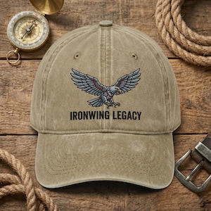 May include: A khaki baseball cap with the text "IRONWING LEGACY" below an eagle graphic with outstretched wings. The eagle is gray and red. The cap is on a wooden surface with a compass and rope.