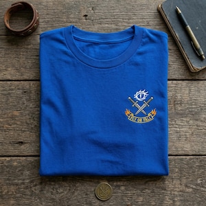 以下が含まれることがあります： クルーネックのロイヤルブルーTシャツ。シャツには、交差した剣、目、そして黄色で「FLY OR FALL」と書かれた小さなエンブレムが付いています。革のブレスレット、ノート、ペンが背景に見えます。