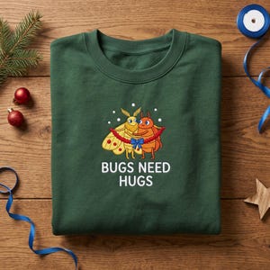 May include: A dark green crewneck sweatshirt featuring a graphic of a yellow moth and an orange beetle hugging, wrapped in a red garland with a blue bow. The text BUGS NEED HUGS is printed in white below the illustration of the two insects.