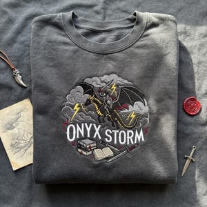 May include: Gray sweatshirt with an embroidered design featuring a black dragon ridden by a knight, with the text "ONYX STORM". The design includes lightning bolts, books, and swords. The sweatshirt is folded.