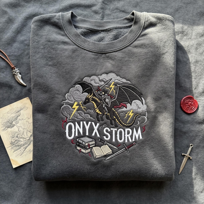 May include: Gray sweatshirt with an embroidered design featuring a black dragon ridden by a knight, with the text "ONYX STORM". The design includes lightning bolts, books, and swords. The sweatshirt is folded.