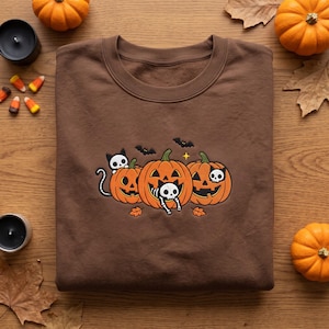 May include: A brown crewneck sweatshirt featuring a central graphic of three orange jack-o'-lanterns. The design includes two black and white skeleton cats, two small black bats, and a yellow star, all arranged in a festive Halloween scene.