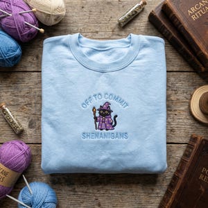 May include: Pale blue sweatshirt featuring embroidered text and a cartoon cat wizard. The text says "OFF TO COMMIT SHENANIGANS". The cat is dressed in a purple wizard outfit and holds a staff. The sweatshirt is made of a soft fabric.