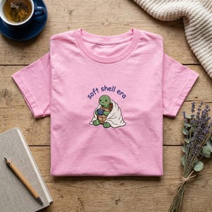 以下が含まれることがあります： 「soft shell era」の文字と刺繍されたカメのデザインが施された、明るいピンク色のTシャツ。カメは毛布に包まれ、カップを持っています。Tシャツは木製の表面に折りたたまれています。
