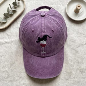 May include: A purple baseball cap with a black cat embroidered on a wine glass. The cap has a curved brim and an adjustable strap. The cat is arched, as if jumping. The cap is made of a soft, washed fabric.