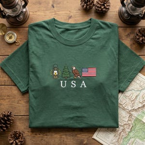 May include: Green t-shirt with embroidered USA-themed design. Features a lantern, pine tree, eagle, and American flag. The word "USA" is embroidered below the images. The shirt is displayed on a wooden surface.