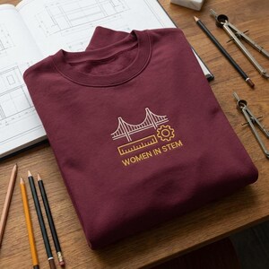May include: A maroon sweatshirt with a crew neck. It has a gold and white graphic of a bridge, gear, and ruler, with the text "WOMEN IN STEM". Pencils, a compass, and a drafting book are on the wooden surface.