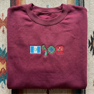 May include: A maroon sweatshirt with embroidered designs. The designs include the Guatemalan flag, a colourful bird, and geometric shapes. The sweatshirt is folded neatly on a patterned surface.