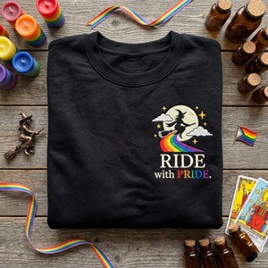 May include: A black crewneck sweatshirt featuring an embroidered graphic of a witch flying on a broomstick across a full moon, leaving a rainbow trail. Below the graphic, the text reads RIDE with PRIDE in white and rainbow-colored lettering.