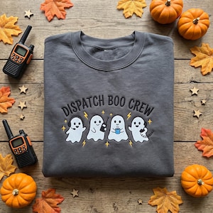 Peut inclure: Un sweat-shirt gris foncé à col rond avec une broderie représentant quatre fantômes blancs et le texte DISPATCH BOO CREW. Les fantômes sont illustrés avec un casque, une radio portable, une tasse de café bleue et un combiné téléphonique, avec des étoiles jaunes.