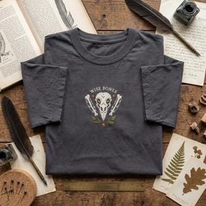 May include: A charcoal gray t-shirt featuring an embroidered design of a bird skull centered above the text "WISE BONES". The graphic includes two feathers flanking the skull, small orange star accents, and green leaves at the base of the design.