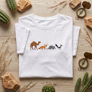 Peut inclure: Un t-shirt blanc à manches courtes orné d'une rangée horizontale d'animaux du désert brodés. Le motif comprend un chameau marron, un fennec orange, un tatou gris et un grand géocoucou bleu et blanc centré sur la poitrine.