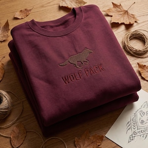 May include: A folded burgundy sweatshirt featuring the embroidered words "WOLF PACK" and a running wolf. The garment is displayed on a wooden surface, complemented by autumn leaves and a coil of twine.