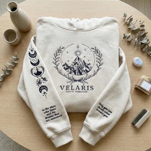 May include: A cream-coloured hoodie featuring a mountain and sun graphic, the word "VELARIS" and the text "CITY OF STARLIGHT". The left sleeve displays moon phases and the text "to the stars who listen and the dreams that are answered". The right sleeve has the text "Only you can decide what breaks you".