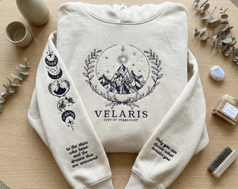 May include: Cream-colored hoodie with a mountain and sun graphic, the word "VELARIS" and the text "CITY OF STARLIGHT". The left sleeve features moon phases and text that reads "to the stars who listen and the dreams that are answered". The right sleeve has the text "Only you can decide what breaks you".
