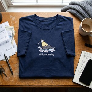 May include: A navy blue t-shirt featuring a graphic of a gray cat lying on a pile of papers under a desk lamp, with the text "still processing" printed in white below the illustration. The design is centered on the chest of the garment.
