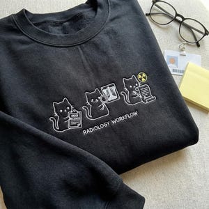May include: Black sweatshirt with white embroidered cat illustrations depicting a radiology workflow. The cats are shown with an order, an X-ray, and a document with a radiation symbol. The text "RADIOLOGY WORKFLOW" is below the cats.