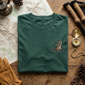 May include: A dark green t-shirt featuring an embroidered raccoon with a backpack. The raccoon is walking on a trail. The shirt is displayed on a wooden surface with a map, compass, hiking poles, and other outdoor gear.