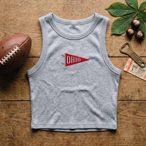 May include: A light gray ribbed tank top featuring a small embroidered red pennant patch with the white text OHIO centered on the chest. The garment has a round neckline and sleeveless design, showcasing a textured fabric construction.