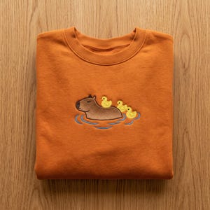 May include: A burnt orange crewneck sweatshirt featuring a central embroidered design of a brown capybara swimming in blue water. Three small yellow ducklings are positioned on the back of the capybara, creating a detailed graphic.