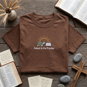 以下が含まれることがあります： 茶色のTシャツ。カメ、開いた本、太陽のグラフィックが描かれています。「Patient in the Promise」の文字が下にあります。木製の表面に折りたたまれたTシャツです。