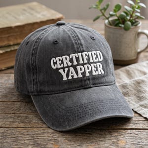 Peut inclure: Une casquette de baseball gris anthracite avec une construction à six panneaux et une visière incurvée. L'avant de la casquette présente le texte "CERTIFIED YAPPER" brodé en lettres majuscules blanches, grasses et sans empattement.