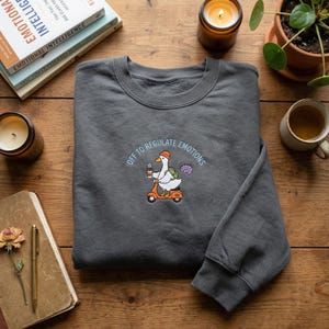 Peut inclure: Sweat-shirt gris foncé avec la phrase brodée "OFF TO REGULATE EMOTIONS". Le motif représente une oie sur un scooter orange, tenant un café et un cerveau. Le sweat-shirt est posé sur une surface en bois avec des livres, des bougies et une tasse.