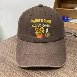May include: Brown baseball cap with the embroidered text "GARDEN HAIR don't care." The cap features a design of sunflowers in a pot, a trowel, and a gardening glove. The cap has a curved peak and a button on top.