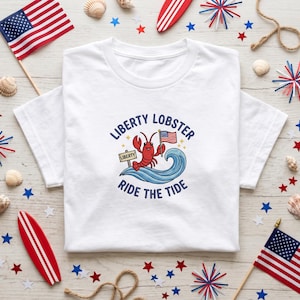May include: A white short-sleeved t-shirt featuring a graphic of a red lobster riding a blue wave. The lobster holds a small American flag and stands next to a sign that reads LIBERTY. The text LIBERTY LOBSTER RIDE THE TIDE surrounds the graphic.