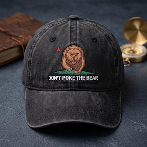 May include: A charcoal grey washed baseball cap featuring an embroidered brown bear standing on a green patch of grass with a small red star above it. The text DON'T POKE THE BEAR is embroidered in white capital letters below the bear graphic.