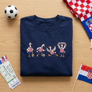 May include: A navy blue sweatshirt featuring an embroidered graphic of four white geese playing soccer. The geese wear red and white checkered jerseys and blue shorts. One goose holds a banner that reads "CROATIA" above its head.