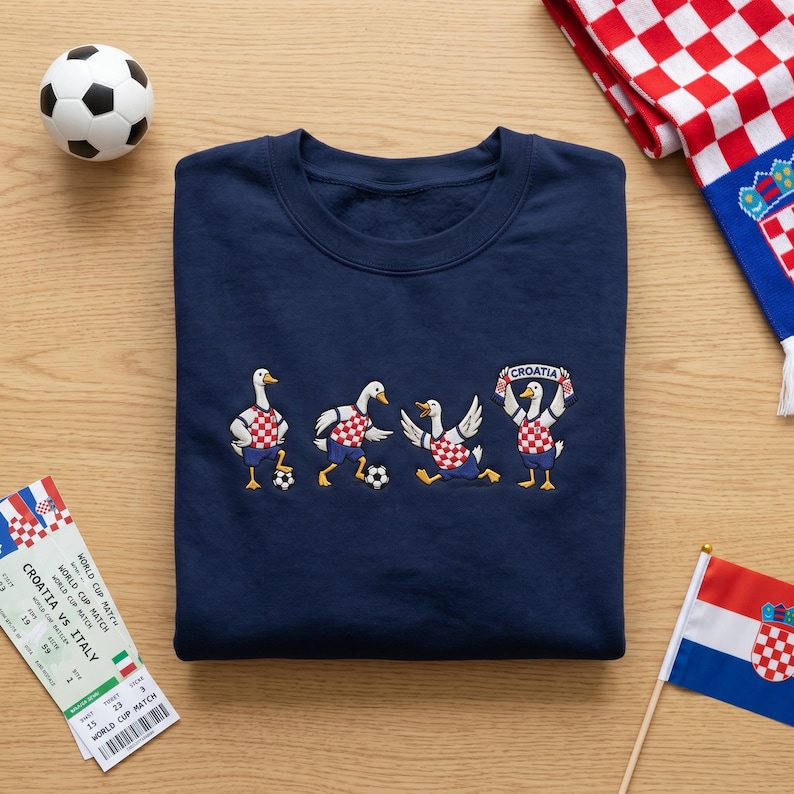 May include: A navy blue sweatshirt featuring an embroidered graphic of four white geese playing soccer. The geese wear red and white checkered jerseys and blue shorts. One goose holds a banner that reads "CROATIA" above its head.