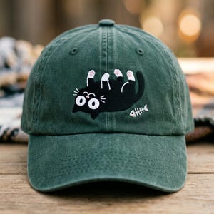 May include: A dark green baseball cap featuring an embroidered design of a black cat lying on its back with white paws and large eyes. Next to the cat is a small white embroidered fish skeleton. The cap has a curved brim and a button on top.