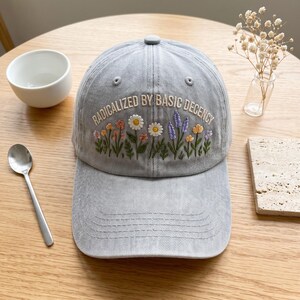 May include: A light grey baseball cap featuring embroidered wildflowers in shades of yellow, orange, purple, and green. The text RADICALIZED BY BASIC DECENCY is stitched in white thread above the floral design. The cap has a curved brim and eyelets.
