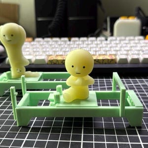 Może przedstawiać: Dwie ż&oacute;łte figurki, jedna siedząca ze złączonymi dłońmi, druga stojąca do g&oacute;ry nogami. Obie znajdują się na jasnozielonych miniaturowych ł&oacute;żkach. Tło obejmuje klawiaturę i czarno-białą kratkę.