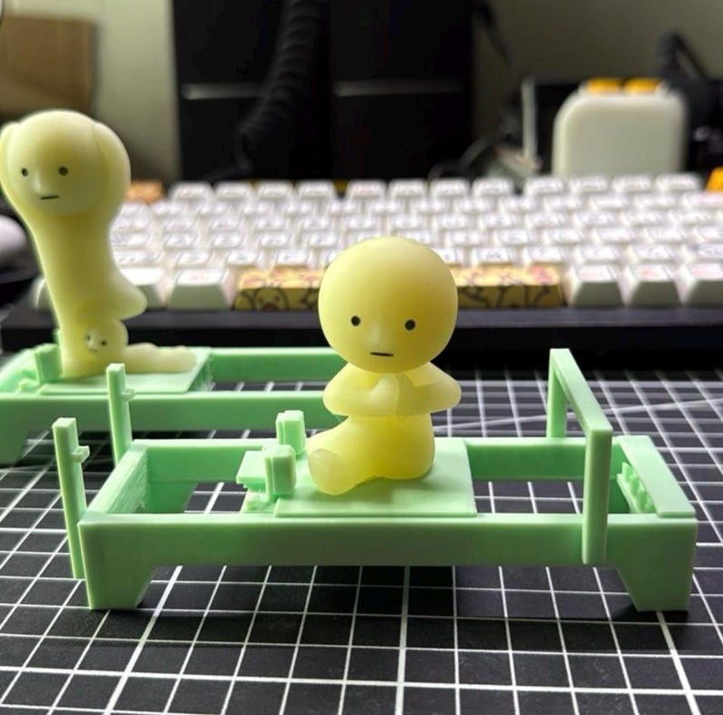 Może przedstawiać: Dwie ż&oacute;łte figurki, jedna siedząca ze złączonymi dłońmi, druga stojąca do g&oacute;ry nogami. Obie znajdują się na jasnozielonych miniaturowych ł&oacute;żkach. Tło obejmuje klawiaturę i czarno-białą kratkę.