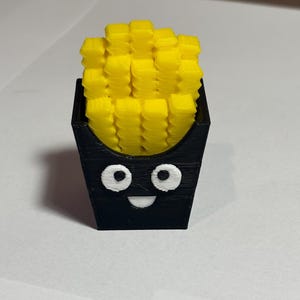 May include: A 3D-printed novelty item featuring a black container with a smiling face, filled with yellow french fries. The fries are stacked neatly, and the container has white eyes and a curved mouth.