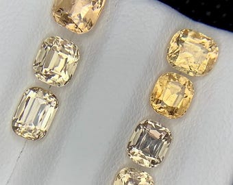 May include: Eight faceted gemstones in shades of yellow and gold. The gems are cushion and emerald cuts, with a brilliant, reflective surface. The stones are arranged on a white surface, with a dark background.