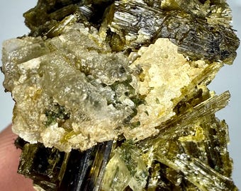 Może przedstawiać: Zbliżenie okazu mineralnego z skupiskiem ciemnozielonych, wydłużonych kryształów. Kryształy są przeplatane jaśniejszymi, przezroczystymi formacjami i kremową, białawą matrycą. Ogólna tekstura jest szorstka i krystaliczna.