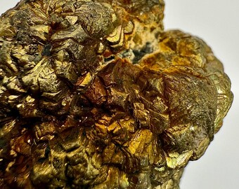 Może przedstawiać: Zbliżenie złotego okazu mineralnego o krystalicznej strukturze. Powierzchnia ma metaliczny połysk i teksturowany wygląd, z odcieniami złota i brązu. Minerał wydaje się być skupiskiem zazębiających się kryształów.