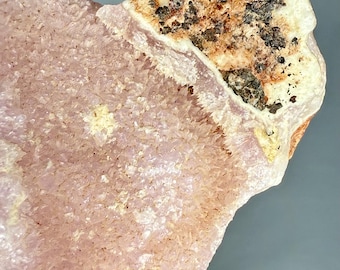 May include: A close-up of a raw, uncut mineral specimen. The stone features a light pink, crystalline structure with a rough, textured surface. The top portion has a beige and brown coloration with dark flecks.