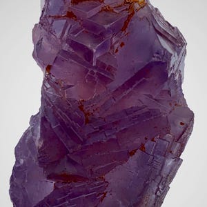 May include: A close-up of a purple fluorite crystal with a layered, geometric structure. The crystal has a translucent quality, with areas of deep purple and reddish-brown inclusions. The overall shape is irregular and natural, showcasing the mineral's unique formation.