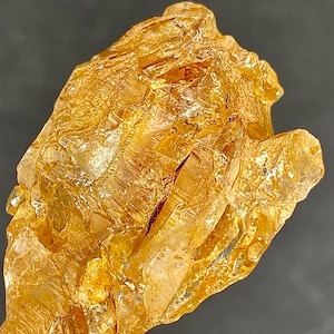 Op de afbeelding: Een close-up van een goudgeel kristalcluster met een ruw, getextureerd oppervlak. Het kristal heeft meerdere facetten en een doorschijnend uiterlijk, met licht dat weerkaatst op de oppervlakken. De achtergrond is donkergrijs.