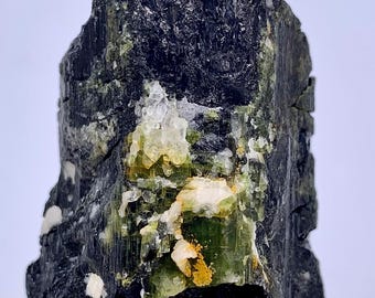 May include: A natural black tourmaline crystal with green and white inclusions. The rough gemstone has a textured surface and visible crystalline structures. Small patches of yellow and clear quartz are also present.
