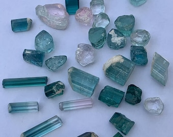 May include: A collection of raw, uncut gemstones in various shades of blue, green, and pink. The stones vary in shape and size, with some appearing crystalline and others more rounded. The gemstones are scattered on a white surface.